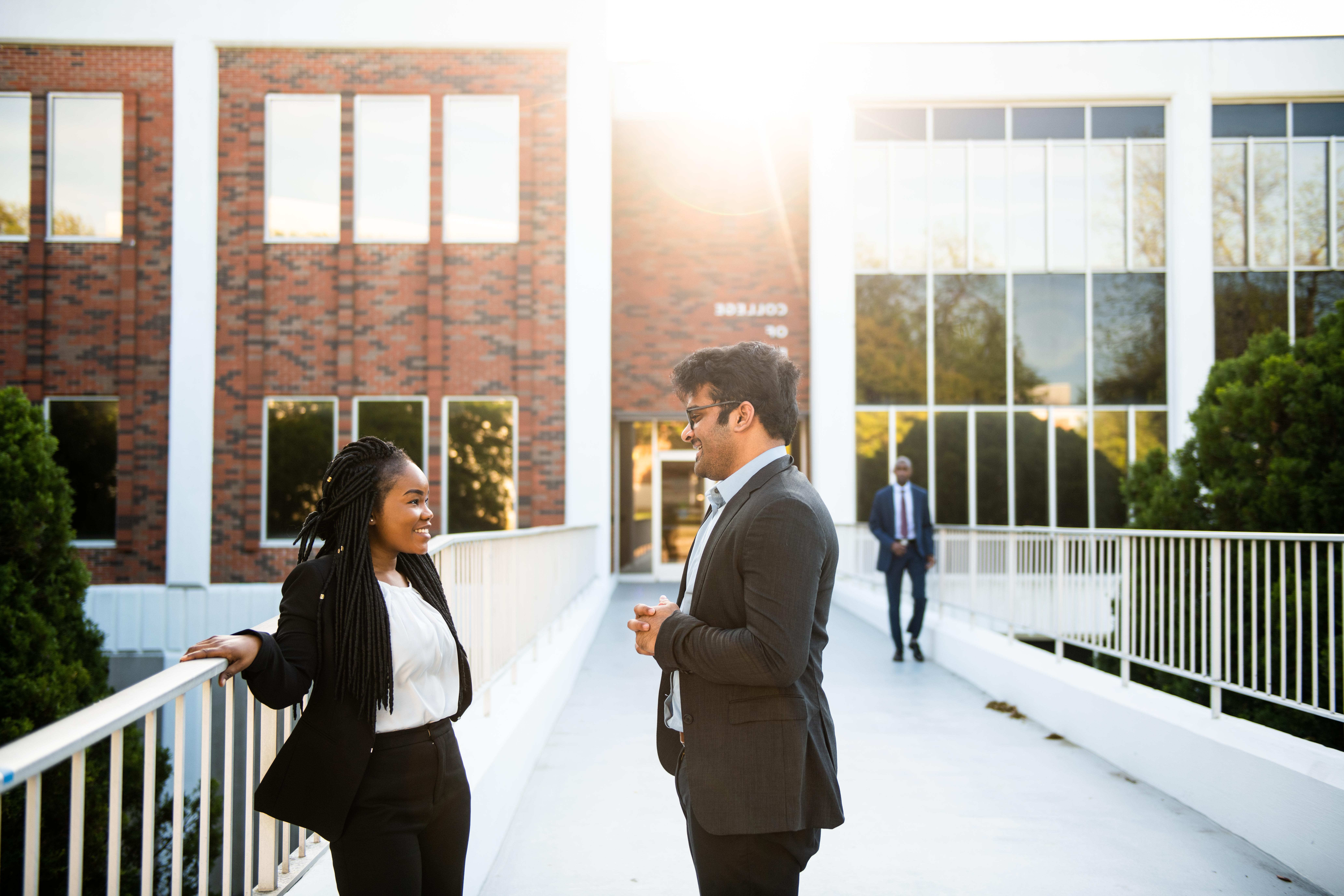 A man and a woman, both wearing business attire, engaged in conversation while standing on a walkway in front of a brick building in the evening light.